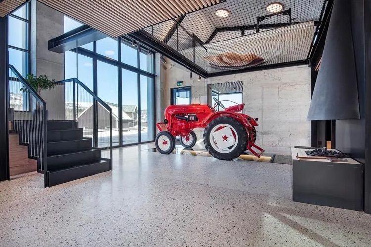 En rød traktor står i en moderne stue med store vinduer og en trapp i bakgrunnen.