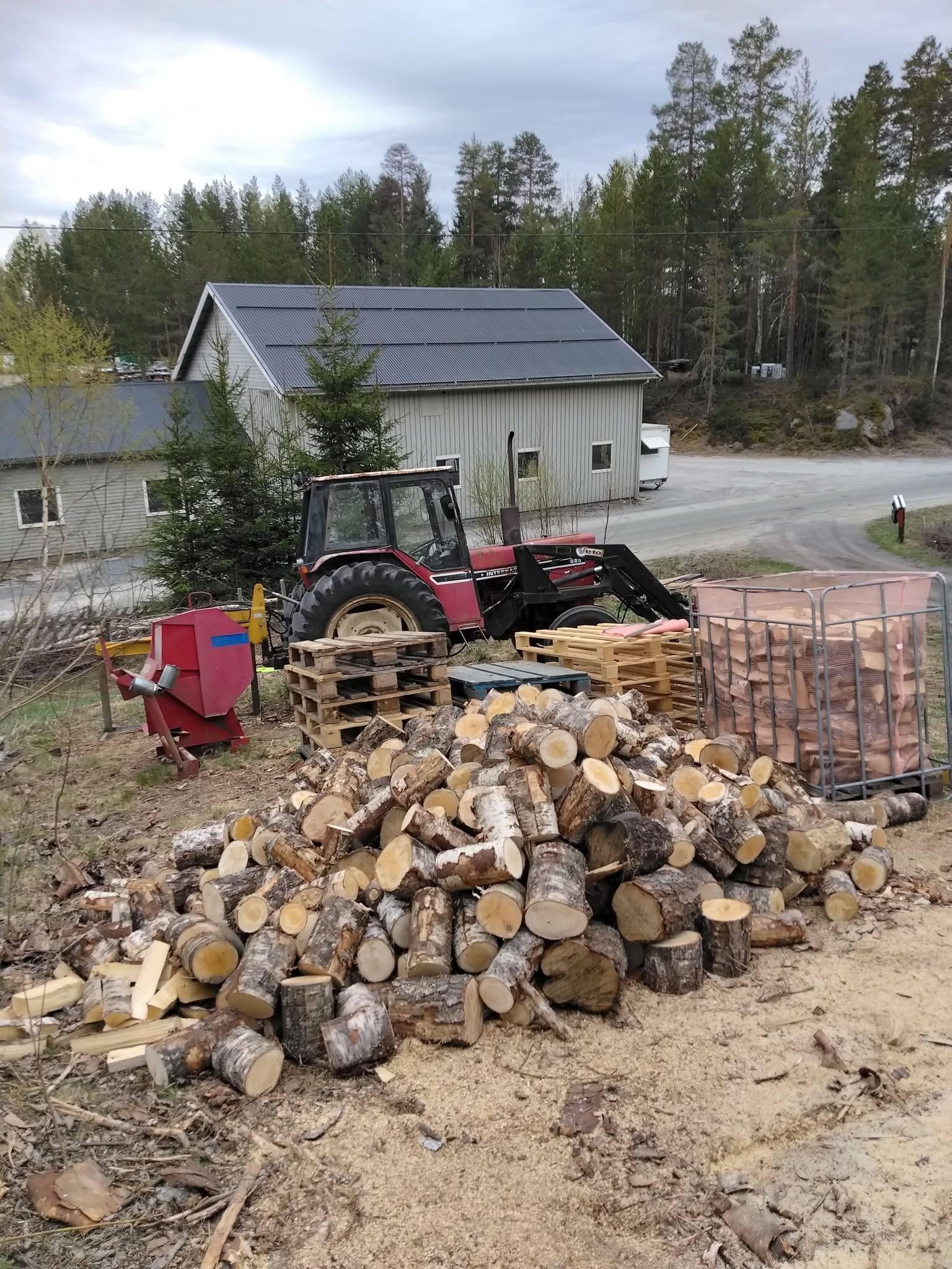 En traktor står ved siden av en haug med ved, med en bygning og en vei i bakgrunnen.