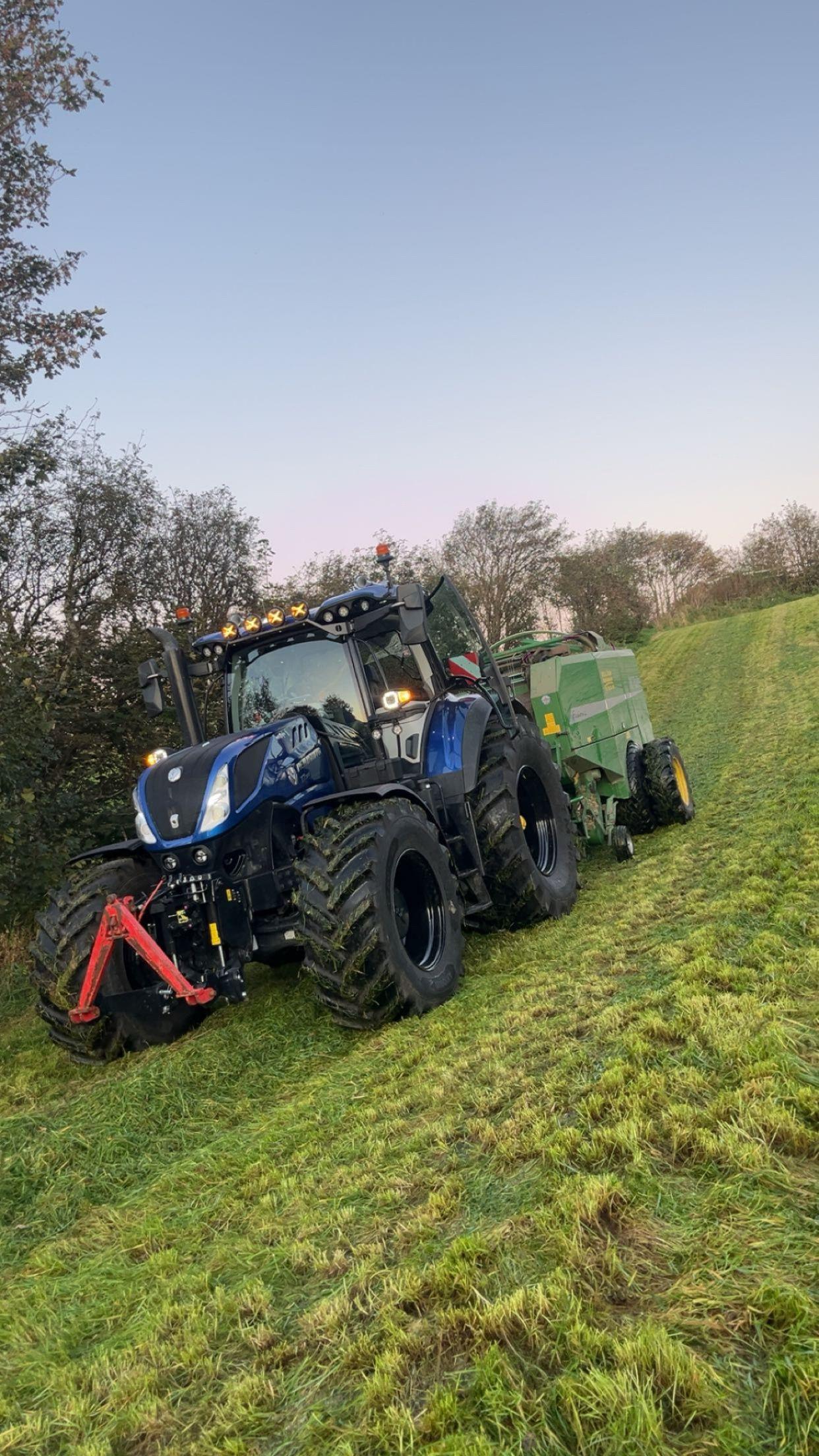 En blå traktor med stor bakhjul og en grønn tilhenger på en grønn eng under en klar himmel.