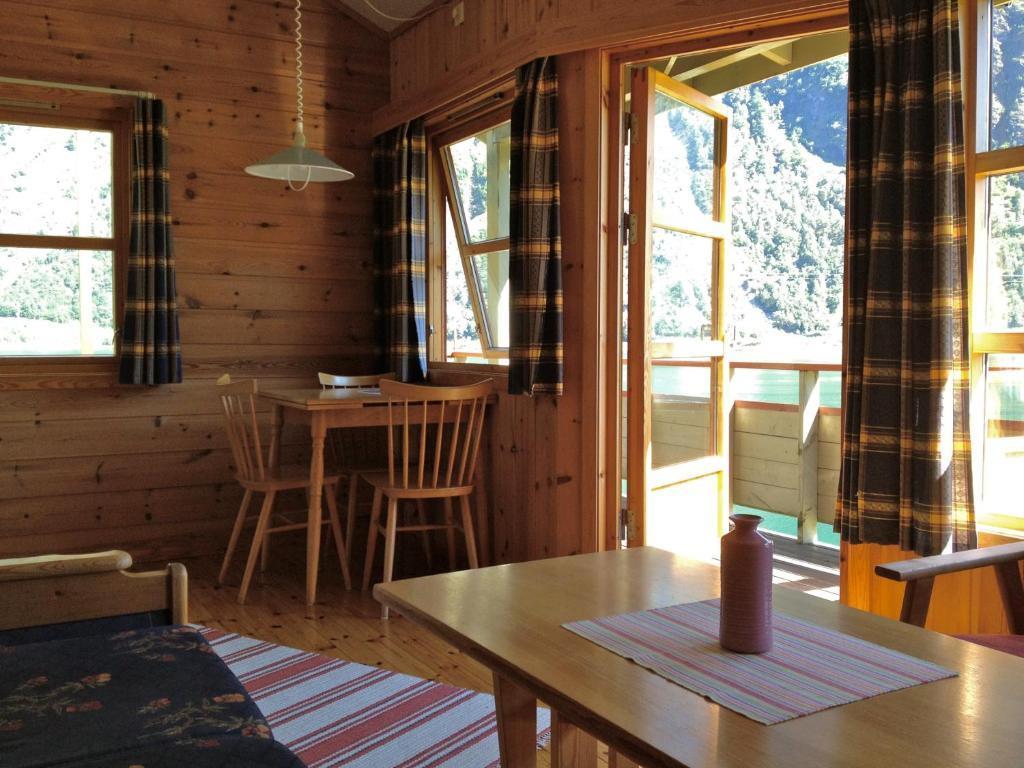 Koselig stue i en hytte med trevegger, bord, stoler og utsikt mot fjellene gjennom åpne vinduer.