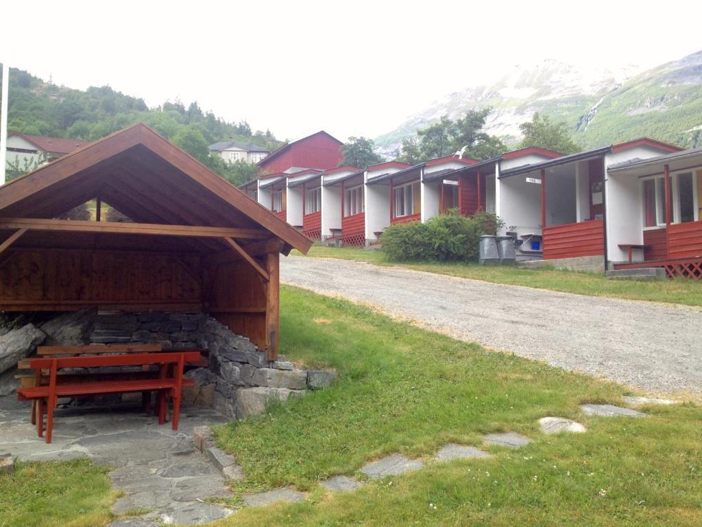 Hytter med rød fasade og en sittegruppe under tak i en grønn hage med fjell i bakgrunnen.