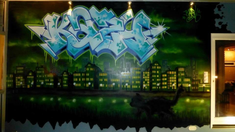 Graffiti med teksten "KAZE" i blått på en vegg, med en mørk bysilhuett og grønne nyanser i bakgrunnen.