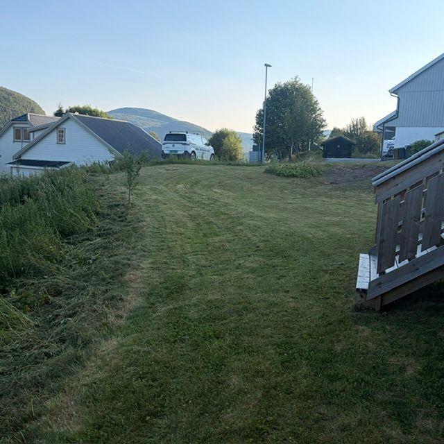 En grønn plen med klippet gress, hus på begge sider og fjell i bakgrunnen under en klar himmel.