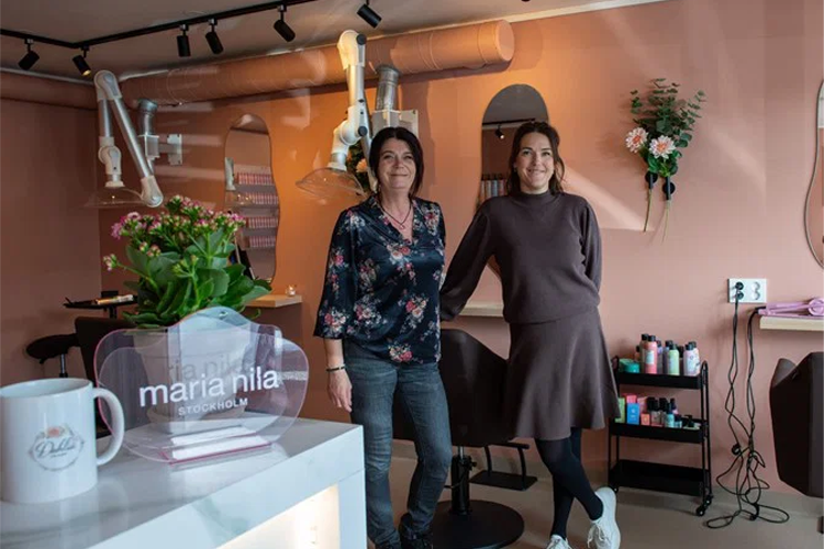 To kvinner står i en frisørsalong med blomster og produkter i bakgrunnen.