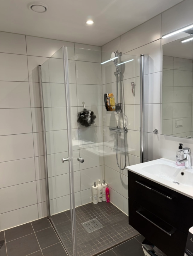 Moderne baderom med dusj, speil, servant og dusjprodukter på hyllen. Fliser i lyse farger.
