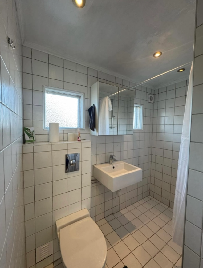 Lyst baderom med fliser, dusj, servant, toalett og speil, godt opplyst av taklamper.