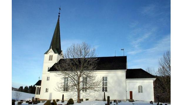 En hvit kirke med spire, omgitt av snø og gravsteiner under en klar blå himmel.