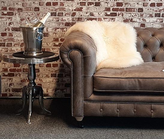 En brun chesterfield-sofa med en hvit pelspledd og et bord med en champagnekjøler i et rom med murvegg.