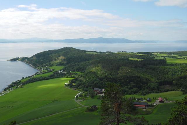 En panoramautsikt over frodige grønne åser og innsjøen, med fjell i bakgrunnen og spredte gårder.