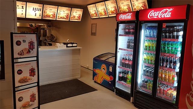 Kiosk med menyer, kjøleskap med Coca-Cola og brus, samt en fryser med snacks og en disk for bestilling.