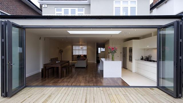 Moderne stue med åpne dører, lys innredning og et kjøkken med hvite skap og spisebord i tre.