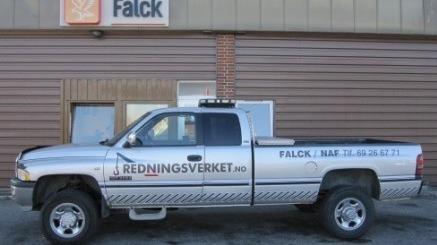 En sølvfarget redningsbil med teksten "Redningsverket" og "Falck" på siden, parkert foran en bygning.
