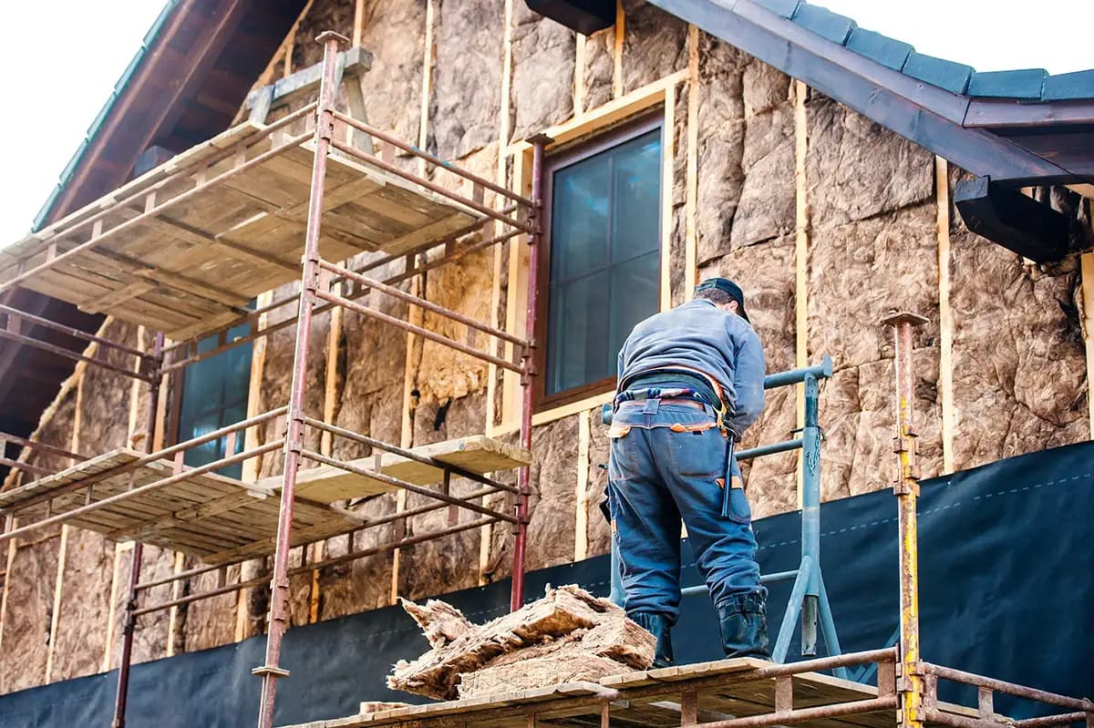 En bygningsarbeider står på stillas og installerer isolasjon på ytterveggen av et hus under renovering.