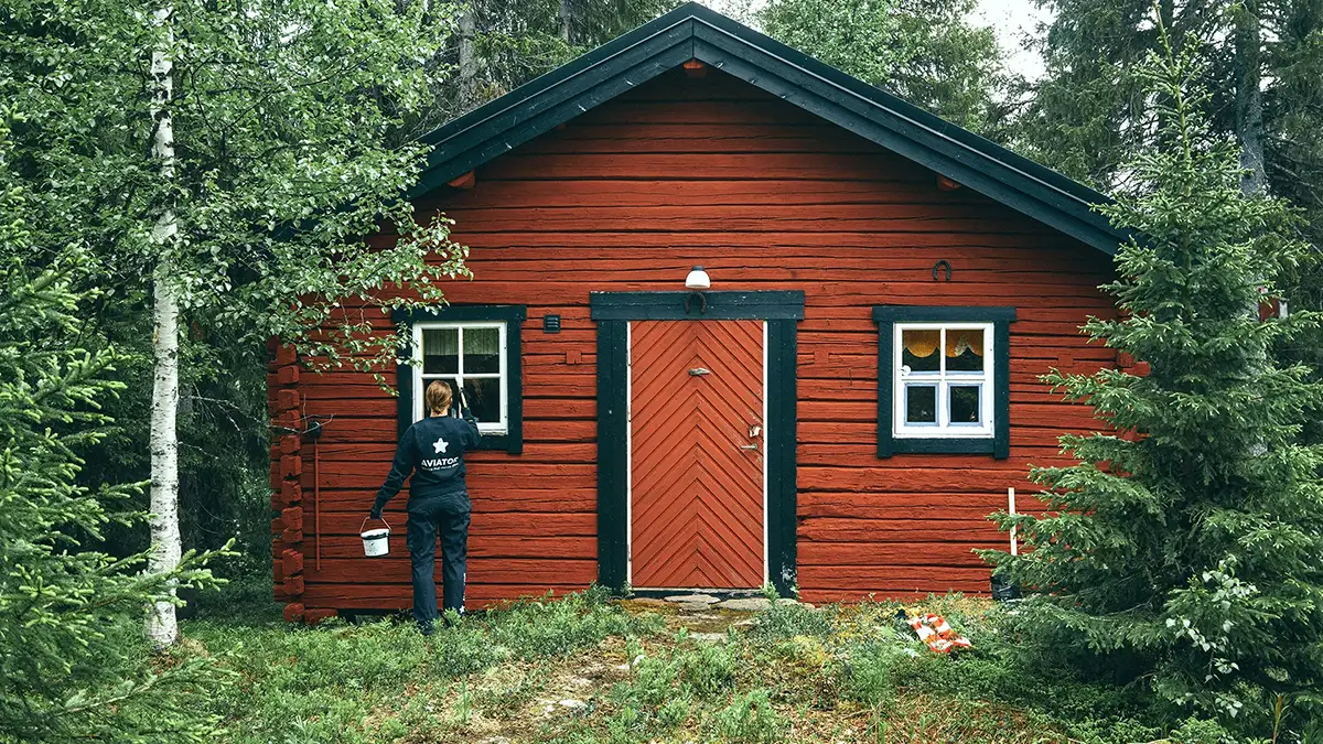 En person som holder en bøtte står foran et rødt hus omgitt av gress og trær.