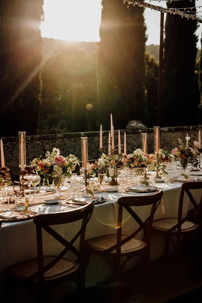 Et utendørs spisebord dekket med blomsterarrangementer, høye lys, tallerkener og glass, omgitt av trestoler ved solnedgang.